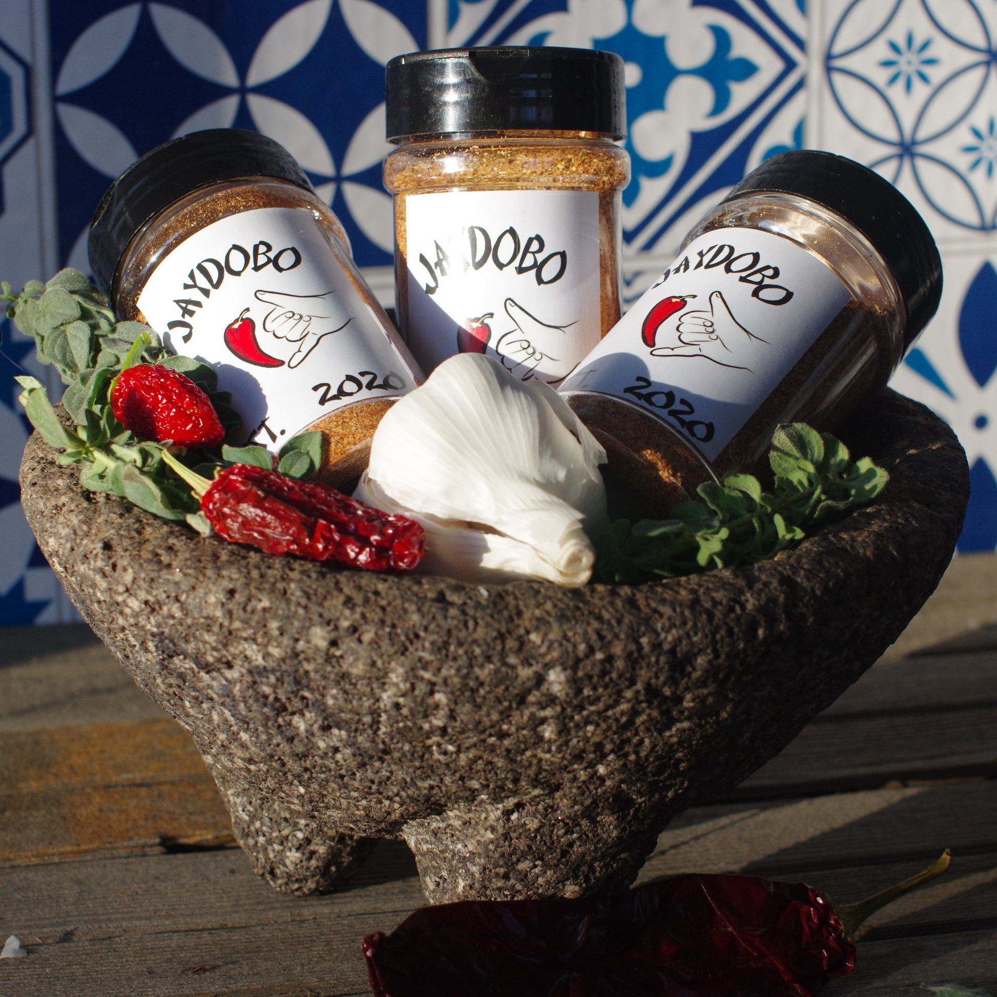 Three Jaydobo spice blend bottles set in a molcajete with oregano, carolina reaper peppers, and garlic bulbs.