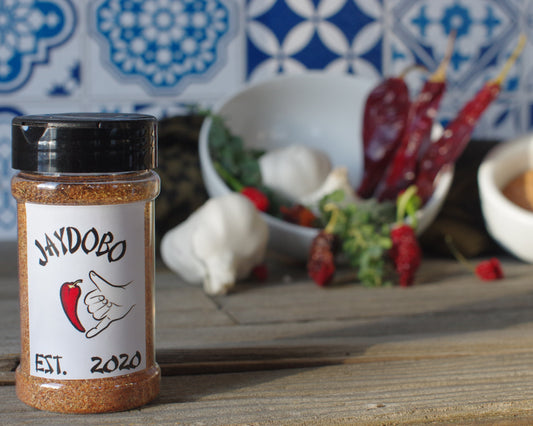 A bottle of Original Jaydobo Spice Blend on a wooden surface with garlic and dried chiles, set against a colorful tiled background.