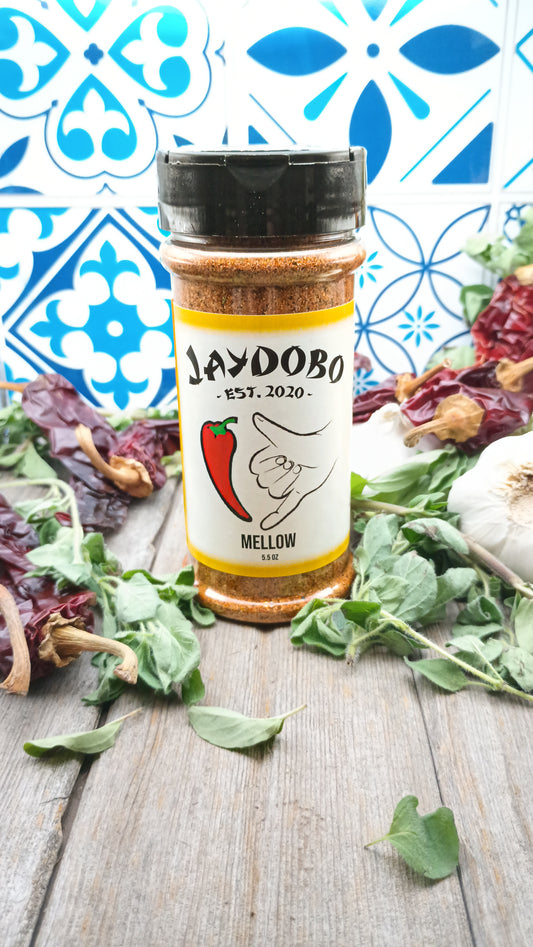 A bottle of Mellow Jaydobo Spice Blend on a wooden cutting board with garlic and dried chiles, set against a colorful tiled background.