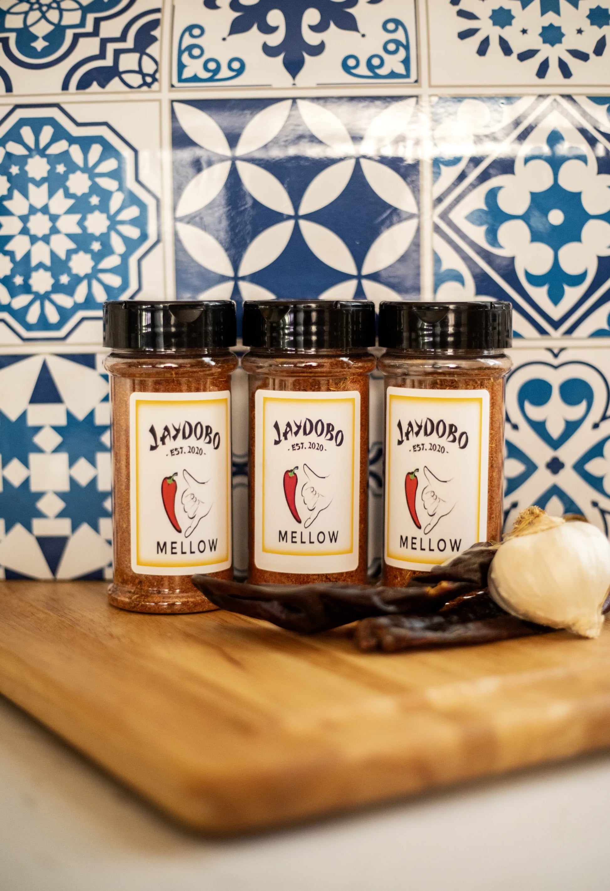  A bundle of three Mellow Jaydobo Spice Blend bottles on a wooden surface with garlic and dried chiles, set against a colorful tiled background.
